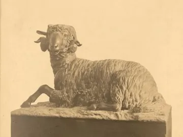 A bronze ram sculpture with curled horns rests on a dark pedestal with a small label near the base.
