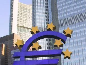 Blue euro sculpture surrounded by twelve yellow stars in front of modern buildings.