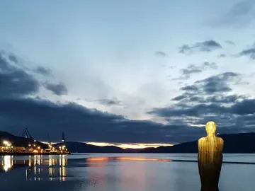 Havmannen sculpture standing in calm water at twilight, with its reflection visible and an illuminated industrial area in the background.