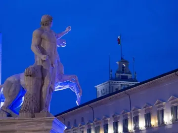 Horse Tamers sculpture lit in blue beside a horse, with an ornate building and clock tower in the background.