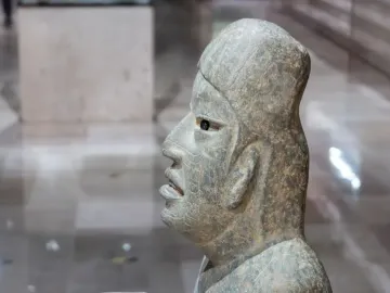 Side profile of the Las Limas Figurine showing a green-grey stone head with an open mouth, visible tongue, and hollow eye socket.
