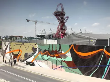 Orbit Tower rises in the background above a brightly tiled mural, with nearby buildings, a stadium, and a crane under a clear sky.
