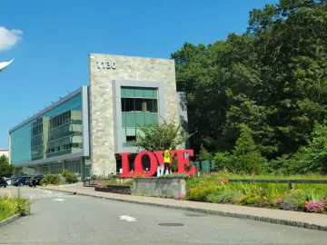Red LOVE sculpture with a tilted O stands in front of a modern stone and glass building, surrounded by flowerbeds and greenery.