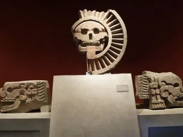 Disk of Mictlāntēcutli displayed with a carved stone skull and flanking stone heads in a museum exhibit.