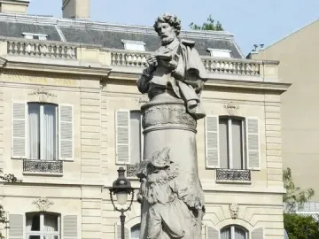 Gavarni Fountain with a stone column topped by a figure holding a book, with relief sculpture and sculpted heads at the base.