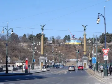 Prague Metronome rising above a tree-lined hillside with a large yellow and blue banner below it.