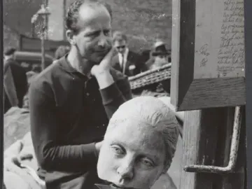 A sculpted female head resting on a metal pitcher, with a man in the background and a framed handwritten sign at right.