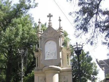 Stone monument from the Monumental Rosary of Montserrat with a central religious relief, a dove, and a cross above it, surrounded by a low fence.