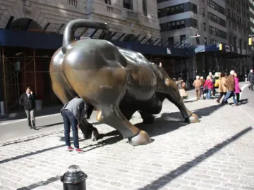 Charging Bull bronze sculpture seen from the rear on a cobblestone street with pedestrians nearby.