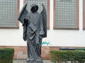 Frankfurter Engel, a dark winged angel statue holding a scroll and a book on a pedestal in a cobblestone plaza.