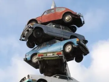 Spindle sculpture with old cars stacked vertically on a central pole, topped by a red Volkswagen Beetle against a blue sky.