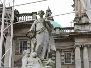 Titanic Memorial statue in Belfast, with a crowned woman holding a wreath above smaller figures and a building with a green domed roof behind it.