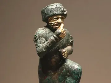 Worshipper of Larsa, a kneeling ancient metal figure with a raised hand, gold-plated face and hands, and a small bowl on its base.