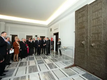 Replica of the Gniezno Doors displayed upright on wooden supports in a bright room with visitors nearby.