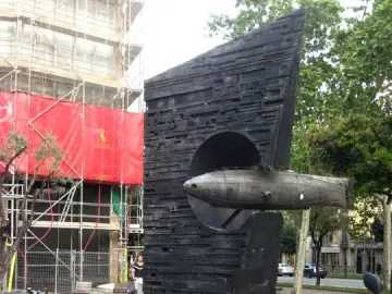 Monument to Narcís Monturiol with a scale submarine model emerging from a dark concrete base, with bicycles in front and a scaffolding-covered building behind.