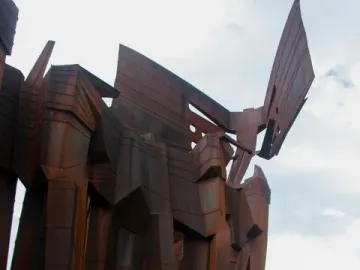 The Monument to the Azoreans, a tall rusted metal sculpture with white graffiti near the base, stands on a grassy area beneath a cloudy sky.