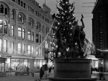 The Three Smiths statue with three muscular figures holding hammers around an anvil in a city square, with a lit Christmas tree behind it.