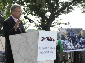 A man speaks at a podium beside a Vietnam Veterans Memorial Fund sign, with a framed display of The Three Soldiers statue on an easel decorated with flowers and flags.