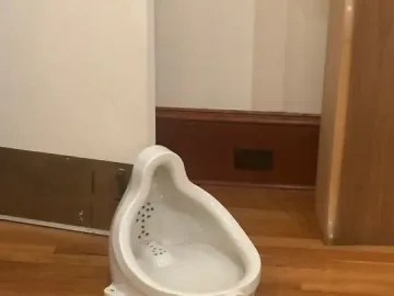A white glazed ceramic urinal sits on a polished wooden floor, with dark dots visible inside and a circular drain opening near the base.