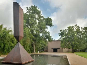 Barnett Newman’s Broken Obelisk sculpture rising from a reflecting pool with trees and a light-toned building behind it.