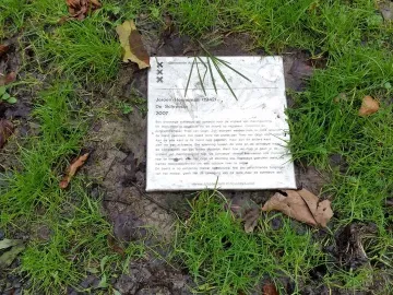 A rectangular plaque for De Schreeuw lies in damp ground among grass and scattered leaves.