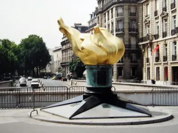 The Flame of Liberty sculpture with a golden flame on a dark stepped base in an urban street setting.