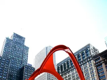 Flamingo, a large red abstract steel sculpture, stands in a city plaza with tall buildings in the background and a person near its base.