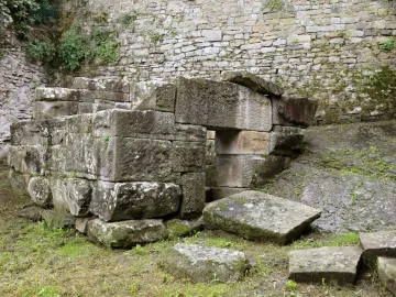 Etruscan tomb built of weathered stone blocks with a small square entrance, set beside a stone wall.
