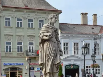 A stone Marian column figure stands behind a wrought-iron fence on a city street.