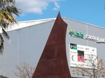 Dorna, a rust-colored abstract metal sculpture with a pointed top and splayed base, set on a stone wall among shrubs.