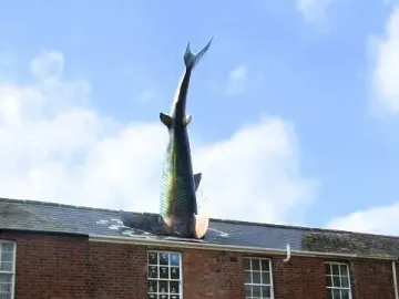 The Headington Shark sculpture plunging into the roof of a red brick building under a blue sky.