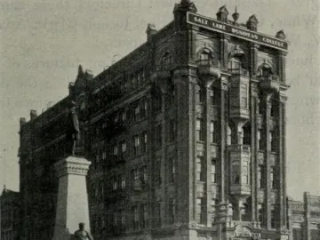 The Brigham Young Monument with a bronze figure atop a tall stone pedestal and relief sculptures at the base, with a white building and bare trees behind it.