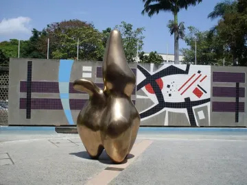 Cloud Shepherd, a smooth golden sculpture, stands on a paved plaza beside a geometric tiled wall.