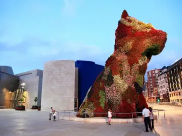 The Puppy flower sculpture stands in an outdoor plaza, covered in red, pink, white, and green flowers, with people gathered nearby.