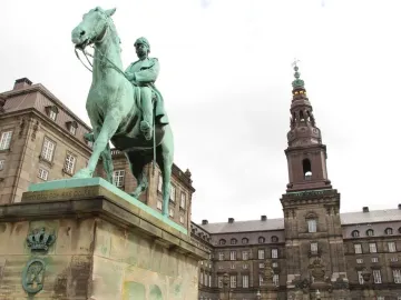 Christian IX equestrian statue on a tall stone pedestal in an open square, with ornate buildings and a slender tower behind it.