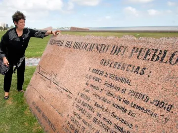 A woman stands beside the Holocaust Memorial plaque, a large angled pink granite monument with engraved text, near a grassy waterfront.