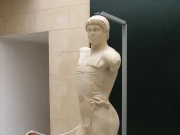 The Motya Charioteer statue displayed on a gray platform in a museum hall.