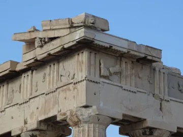 Weathered marble corner of the Parthenon pediments with fluted columns, triglyphs, worn relief metopes, and a lion\'s head against a blue sky.