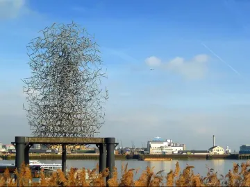 Quantum Cloud sculpture beside the water with buildings on the far shore under a clear sky.