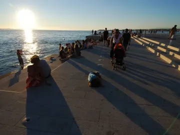People gather along the Sea Organ promenade in Zadar at sunset, with dark circular openings visible in the stone steps by the sea.