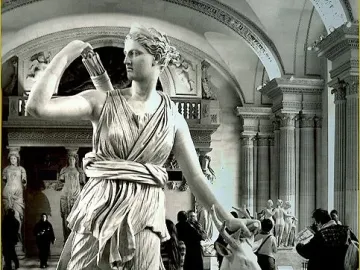 The Diana of Versailles marble statue stands in a museum hall with a small deer beside it and visitors in the background.