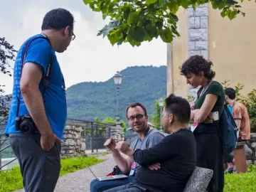 Five people gathered outdoors, with two seated on a stone bench and others standing nearby in conversation.
