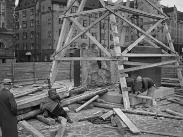 Workers erecting the Aleksis Kivi Memorial within wooden scaffolding beside stone blocks and construction materials.