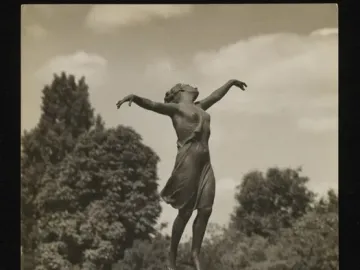 The Dancing Girl sculpture stands on a tall pedestal outdoors among trees.
