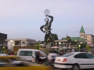 La Nouvelle Liberté towering metal sculpture in a busy street, with a spherical cage held overhead and a church spire in the background.