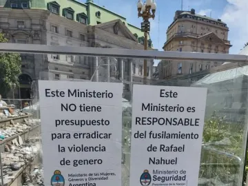 White protest signs attached to a glass barrier at the Manuel Belgrano monument, with rocks and pebbles inside the enclosure.