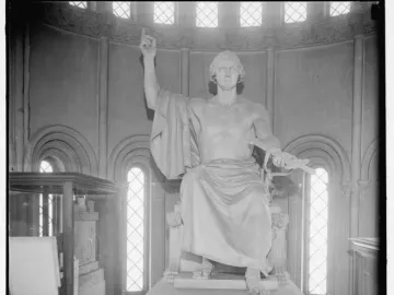 A large seated marble statue of a male figure with one arm raised and the other holding a bound object, displayed in a room with arched windows.