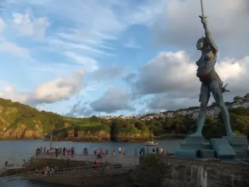 The Verity statue stands beside Ilfracombe pier with water, a grassy hillside, and a cloudy sky in the background.