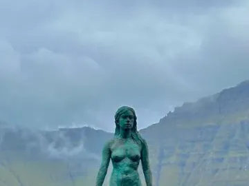 Kópakonan bronze statue standing on dark rocks by the water with misty mountains behind it.