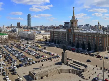 Lenin Monument stands on a tall pedestal in a city square with steps, people, cars, and surrounding buildings.